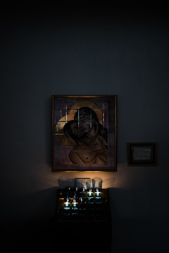 A religious icon of the Virgin Mary holding baby Jesus is mounted on a dimly lit wall. Beneath the icon, several candles are lit, casting a soft, warm glow. There is a small framed plaque on the wall beside the icon.