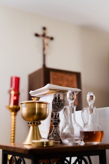 A collection of religious artifacts is displayed on a table, including a golden chalice, a silver candlestick, and glass cruets filled with liquid. In the background, a red candle and a wooden cross on the wall can be seen.