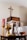 A collection of religious artifacts is displayed on a table, including a golden chalice, a silver candlestick, and glass cruets filled with liquid. In the background, a red candle and a wooden cross on the wall can be seen.