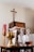A collection of religious artifacts is displayed on a table, including a golden chalice, a silver candlestick, and glass cruets filled with liquid. In the background, a red candle and a wooden cross on the wall can be seen.