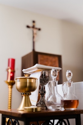 A collection of religious artifacts is displayed on a table, including a golden chalice, a silver candlestick, and glass cruets filled with liquid. In the background, a red candle and a wooden cross on the wall can be seen.