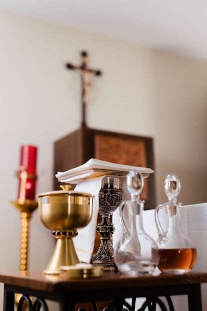A collection of religious artifacts is displayed on a table, including a golden chalice, a silver candlestick, and glass cruets filled with liquid. In the background, a red candle and a wooden cross on the wall can be seen.
