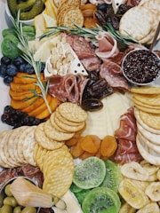 A vibrant charcuterie board featuring an assortment of cured meats, various types of cheese, and several kinds of crackers. Fresh and dried fruits such as kiwi, blueberries, apricots, and dates are artfully arranged alongside olives and green pickles. There is also a small dish containing a dark red jam garnished with a sprig of rosemary.