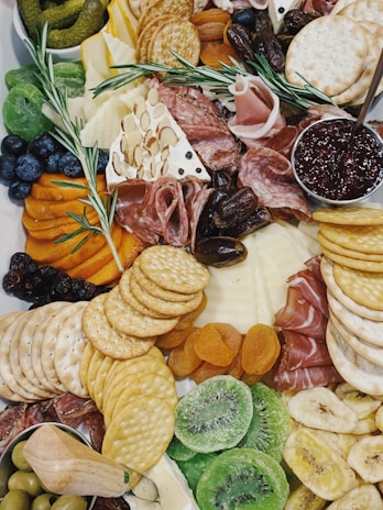 A vibrant charcuterie board featuring an assortment of cured meats, various types of cheese, and several kinds of crackers. Fresh and dried fruits such as kiwi, blueberries, apricots, and dates are artfully arranged alongside olives and green pickles. There is also a small dish containing a dark red jam garnished with a sprig of rosemary.