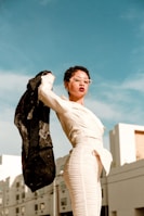 A person in a fashionable white outfit stands confidently against a clear blue sky, holding up a black jacket. The background features modern buildings, adding an urban context to the scene. The person is wearing large hoop earrings and stylish clear-framed glasses.