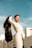 A person in a fashionable white outfit stands confidently against a clear blue sky, holding up a black jacket. The background features modern buildings, adding an urban context to the scene. The person is wearing large hoop earrings and stylish clear-framed glasses.