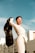 A person in a fashionable white outfit stands confidently against a clear blue sky, holding up a black jacket. The background features modern buildings, adding an urban context to the scene. The person is wearing large hoop earrings and stylish clear-framed glasses.