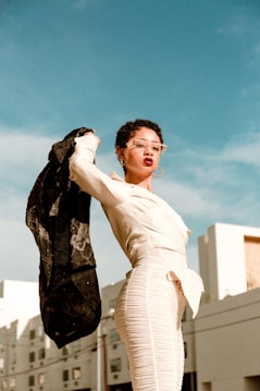 A person in a fashionable white outfit stands confidently against a clear blue sky, holding up a black jacket. The background features modern buildings, adding an urban context to the scene. The person is wearing large hoop earrings and stylish clear-framed glasses.