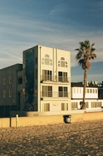 A modern, multi-story beachfront building with large windows and balconies, featuring a mural on its side. A lone palm tree stands nearby, casting its shadow. The sandy beach and a trash can are visible in the foreground, under a clear blue sky.
