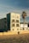 A modern, multi-story beachfront building with large windows and balconies, featuring a mural on its side. A lone palm tree stands nearby, casting its shadow. The sandy beach and a trash can are visible in the foreground, under a clear blue sky.