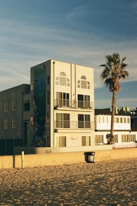 A modern, multi-story beachfront building with large windows and balconies, featuring a mural on its side. A lone palm tree stands nearby, casting its shadow. The sandy beach and a trash can are visible in the foreground, under a clear blue sky.