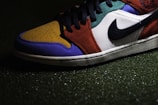 A close-up view of a sneaker with a multicolored design, including sections of mustard yellow, royal blue, burnt orange, and forest green. It features a prominent black Swoosh logo on a white background, and black laces. The shoe is positioned on a textured surface that appears to be artificial grass.