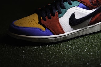 A close-up view of a sneaker with a multicolored design, including sections of mustard yellow, royal blue, burnt orange, and forest green. It features a prominent black Swoosh logo on a white background, and black laces. The shoe is positioned on a textured surface that appears to be artificial grass.