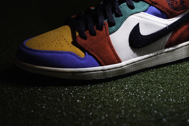A close-up view of a sneaker with a multicolored design, including sections of mustard yellow, royal blue, burnt orange, and forest green. It features a prominent black Swoosh logo on a white background, and black laces. The shoe is positioned on a textured surface that appears to be artificial grass.