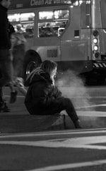 A person is sitting on a curb at night with visible steam or smoke rising nearby. The scene includes a blurred bus in the background with the words 'Clean Air Vehicle' on it, and other pedestrians walking past. The focus is on the solitary figure in the foreground, wearing a heavy-looking coat, suggesting cold weather.