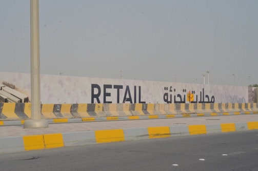 A street scene with a concrete barrier painted in alternating yellow and black stripes. Behind the barrier, a large sign with the word 'RETAIL' and Arabic text is visible, suggesting possible construction or development in the area. The road is mostly empty with clear skies, creating a quiet, urban environment.