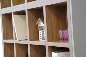 A wooden shelf unit with a grid design featuring a decorative small house in blue and white, several books with colorful spines, and a beige binder. The shelves are made of light wood with multiple compartments.