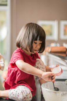 A young child with short dark hair is sitting on a counter, wearing a red shirt and floral-patterned leggings. They are engaged in an activity involving a mixing bowl, spoon, and a small container, appearing focused and absorbed.