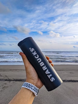 A hand holding a black Starbucks tumbler with white text against a backdrop of a beach with waves and a partly cloudy blue sky.