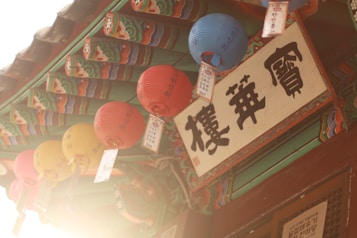Colorful paper lanterns hang from a traditional wooden structure with ornate, painted beams and Korean text on a prominent sign. The sunlight creates a warm, inviting glow.
