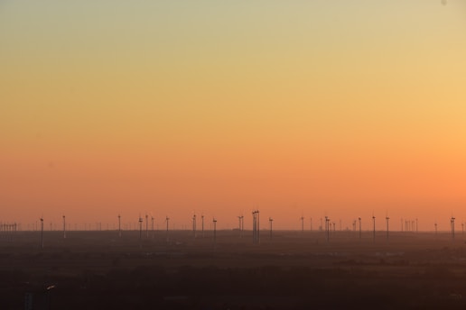 A scenic view of numerous wind turbines spread across a flat landscape during sunset. The sky transitions from a warm orange near the horizon to a pale yellow higher up. The silhouettes of the turbines stand out against the gradient sky, with the land appearing dark in contrast.