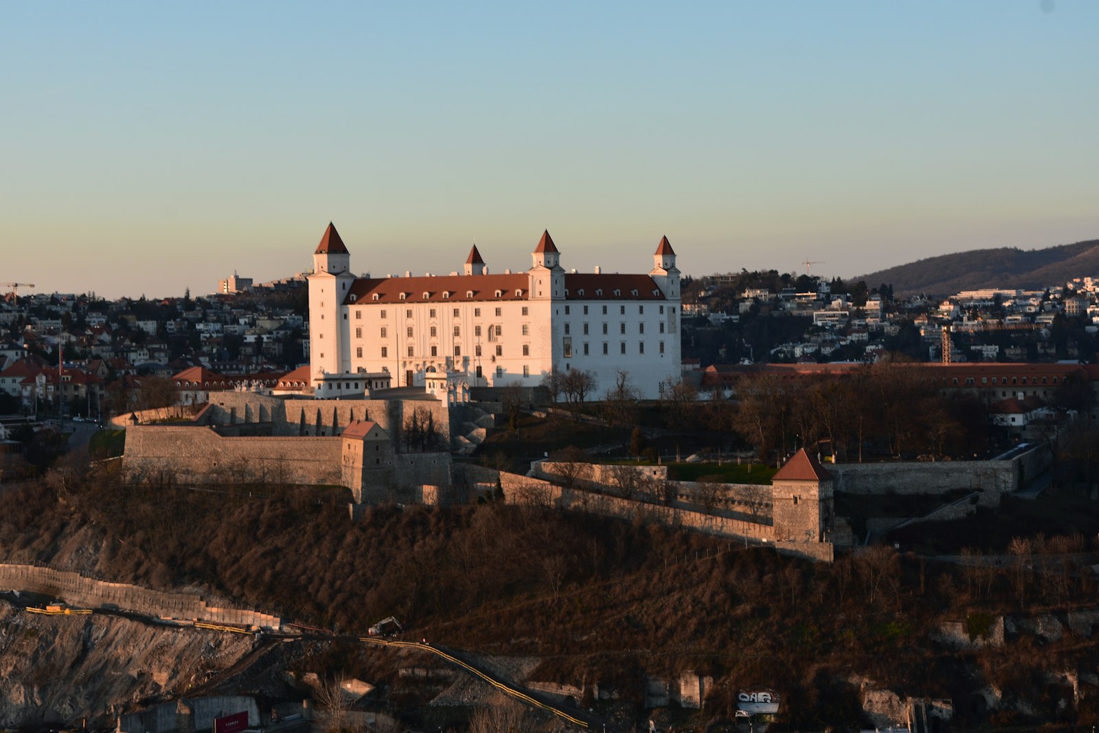 Bratislava Castle's white walls and red-roofed corner towers above the old town in warm sunset light