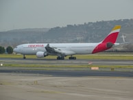 A large commercial airplane is taxiing on the runway at an airport. The aircraft is predominantly white, with red accents on the tail and engines, and the word 'Iberia' is written on the side. The background features a hazy view of a distant hillside and sky.