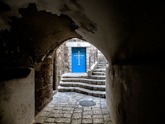 A cobblestone path leads through an arched tunnel into a sunlit area. The focus is on a striking blue door adorned with a cross and Greek letters. Stone steps ascend to the entrance, flanked by rugged stone walls.