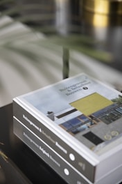 A close-up of two stacked hardcover books with the title 'Buitengewoon & Betaalbaar Bouwen' visible on the spines. The cover displays an image of a modern architectural design featuring a house with a large window and yellow accent panel. The background includes blurred elements, possibly consisting of plant leaves, adding a touch of natural ambiance.