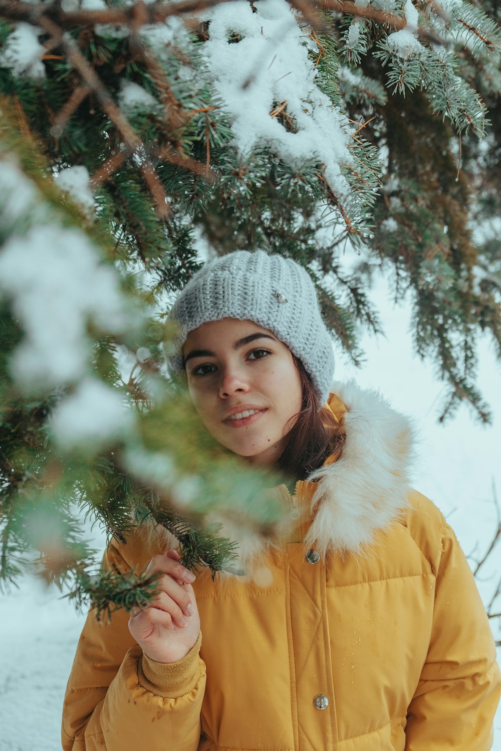 girl in brown jacket and gray knit cap standing near green tree with snow