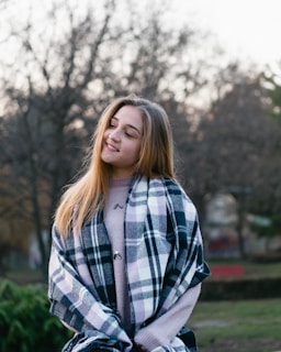 A young woman wearing a soft lavender hoodie with a subtle faith-inspired graphic, smiling warmly in a sunlit city park.