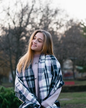 A young woman wearing a soft lavender hoodie with a subtle faith-inspired graphic, smiling warmly in a sunlit city park.