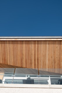 A modern architectural facade featuring vertical wooden panels and clear glass railing under a clear blue sky. The structure includes visible office chairs and a clean, minimalist design.