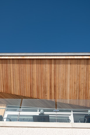 A modern architectural facade featuring vertical wooden panels and clear glass railing under a clear blue sky. The structure includes visible office chairs and a clean, minimalist design.