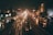 Heavy traffic fills a city street at night, with vehicles lined up on both sides of a central divider. The wet surface of the road reflects the bright headlights and taillights, creating streaks of light. Buildings and street lights illuminate the scene, adding to the urban ambiance.