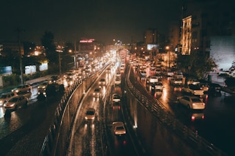 Heavy traffic fills a city street at night, with vehicles lined up on both sides of a central divider. The wet surface of the road reflects the bright headlights and taillights, creating streaks of light. Buildings and street lights illuminate the scene, adding to the urban ambiance.