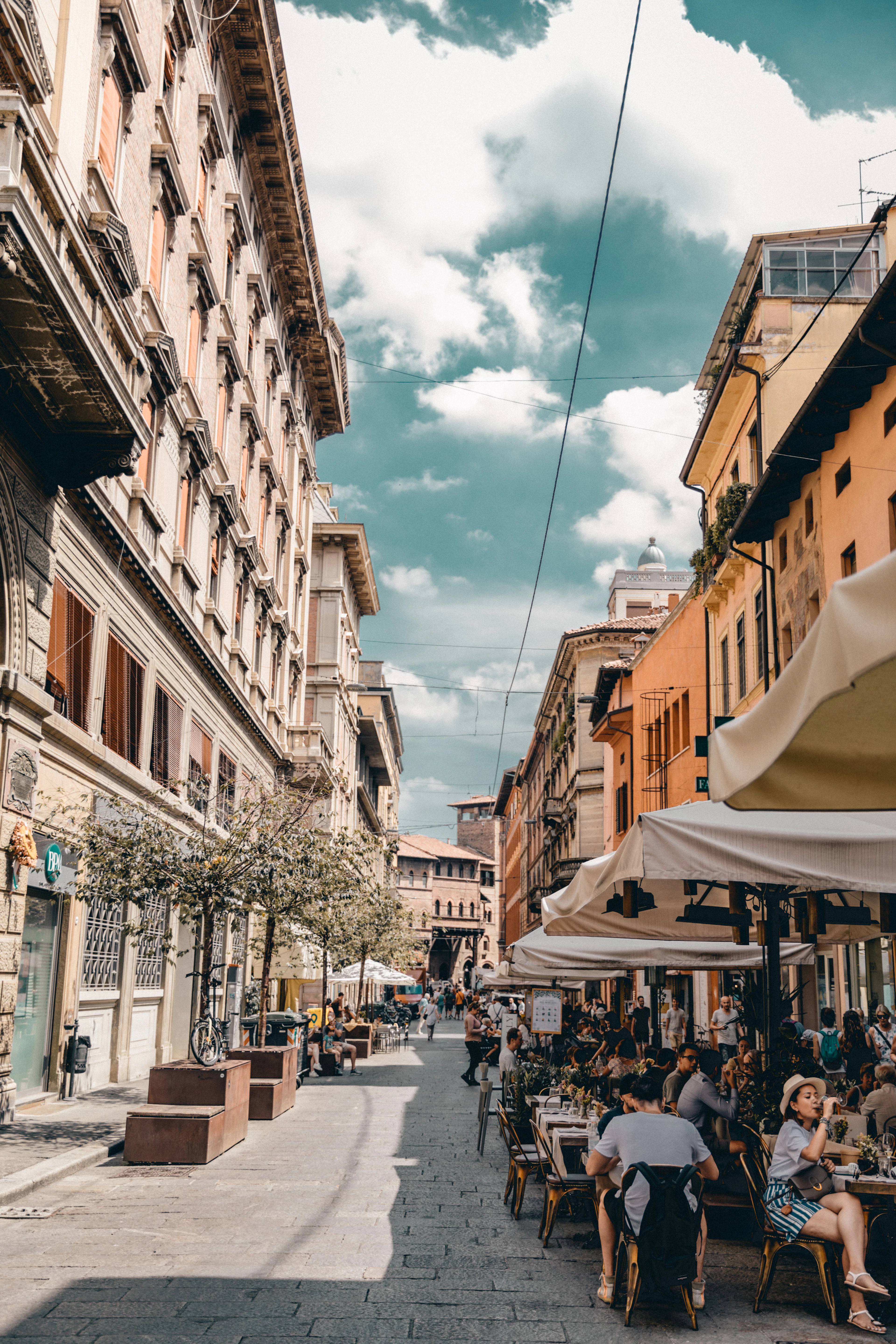 Diners fill outdoor cafe tables along a bustling arcaded Bologna street under a partly cloudy sky.