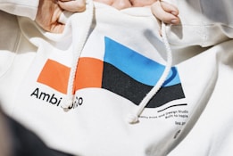 A close-up of a white hooded sweatshirt with a colorful geometric design on the front. The design includes a red and blue rectangle and a black shape. There are textual elements near the design, with legible partial texts such as 'Ambi' and 'gnO'. Two hands are holding the drawstrings of the hoodie.