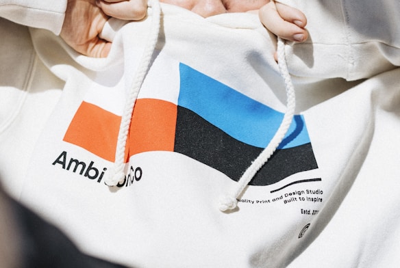 A close-up of a white hooded sweatshirt with a colorful geometric design on the front. The design includes a red and blue rectangle and a black shape. There are textual elements near the design, with legible partial texts such as 'Ambi' and 'gnO'. Two hands are holding the drawstrings of the hoodie.