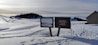 A snowy landscape with gentle hills and tracks leading through the snow. Two wooden signboards are positioned in the foreground, one displaying a picture and the other with text, including contact information for 'Refuge du Semnoz'. In the background, a line of trees marks the horizon, and a small structure stands to the right.