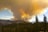 A vast forest landscape with tall trees in the foreground and distant mountains. A large, dense column of smoke rises into the sky, primarily comprising dark and orange hues, indicating a wildfire. The sky is partly cloudy with blue patches visible around the smoke.
