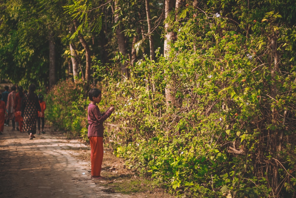 woman in red dress walking on dirt road between green trees during daytime