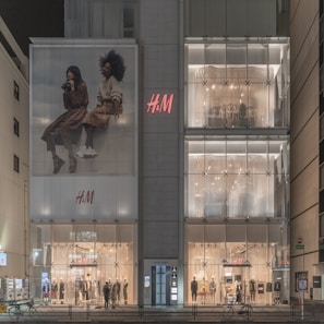 A modern retail store front featuring H&M branding with large glass windows displaying mannequins in fashionable outfits. There is a large advertisement on the building showcasing two individuals in stylish clothing. The store is illuminated, indicating it is evening or nighttime.
