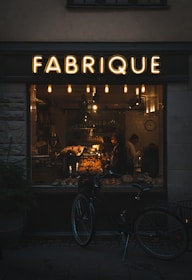 A warmly lit bakery interior can be seen through a large window labeled 'FABRIQUE'. Inside, a variety of baked goods are displayed on shelves and counters, with two people visible. A vintage-style bicycle is parked outside the shop, and several hanging lights inside contribute to a cozy atmosphere.