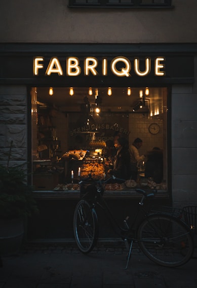 A warmly lit bakery interior can be seen through a large window labeled 'FABRIQUE'. Inside, a variety of baked goods are displayed on shelves and counters, with two people visible. A vintage-style bicycle is parked outside the shop, and several hanging lights inside contribute to a cozy atmosphere.