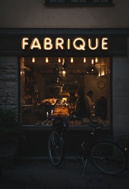 A warmly lit bakery interior can be seen through a large window labeled 'FABRIQUE'. Inside, a variety of baked goods are displayed on shelves and counters, with two people visible. A vintage-style bicycle is parked outside the shop, and several hanging lights inside contribute to a cozy atmosphere.