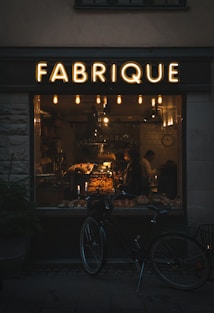 A warmly lit bakery interior can be seen through a large window labeled 'FABRIQUE'. Inside, a variety of baked goods are displayed on shelves and counters, with two people visible. A vintage-style bicycle is parked outside the shop, and several hanging lights inside contribute to a cozy atmosphere.