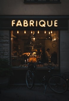 A warmly lit bakery interior can be seen through a large window labeled 'FABRIQUE'. Inside, a variety of baked goods are displayed on shelves and counters, with two people visible. A vintage-style bicycle is parked outside the shop, and several hanging lights inside contribute to a cozy atmosphere.