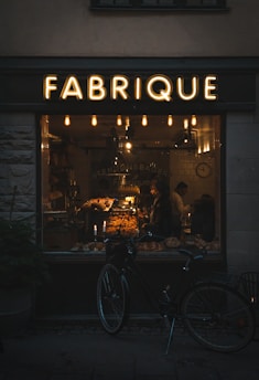 A warmly lit bakery interior can be seen through a large window labeled 'FABRIQUE'. Inside, a variety of baked goods are displayed on shelves and counters, with two people visible. A vintage-style bicycle is parked outside the shop, and several hanging lights inside contribute to a cozy atmosphere.