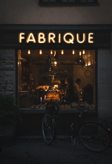 A warmly lit bakery interior can be seen through a large window labeled 'FABRIQUE'. Inside, a variety of baked goods are displayed on shelves and counters, with two people visible. A vintage-style bicycle is parked outside the shop, and several hanging lights inside contribute to a cozy atmosphere.